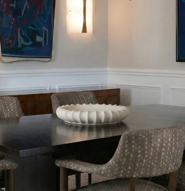 A dining room with a dark table, four patterned chairs, and the Phoebe Bowl 15″ by FlowDecor Inc. as a centerpiece, surrounded by white wainscoting, abstract blue art, and warm wall lighting for a cozy feel.