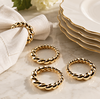 Four L’Objet Jessica McCormack Shell Napkin Ring s/4 - Gold rings with a twisted rope design, one placed around a white cloth napkin and three resting on a marble surface near stacked plates.