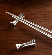 A pair of metal chopsticks resting on two silver-plated Christofle Uni Silver Plated Set of 2 Knife/Chopstick Rests on a wooden surface.