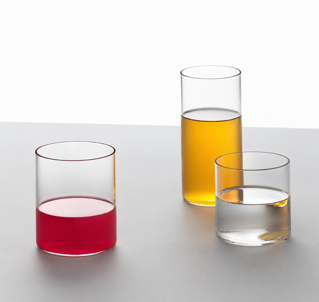 Three Ichendorf Milano Cilindro Water Glass tumblers in hand-blown borosilicate glass on a white surface, filled with red liquid, yellow liquid, and clear water.