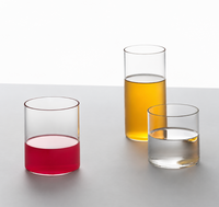 Three Ichendorf Milano Cilindro Water Glass tumblers in hand-blown borosilicate glass on a white surface, filled with red liquid, yellow liquid, and clear water.