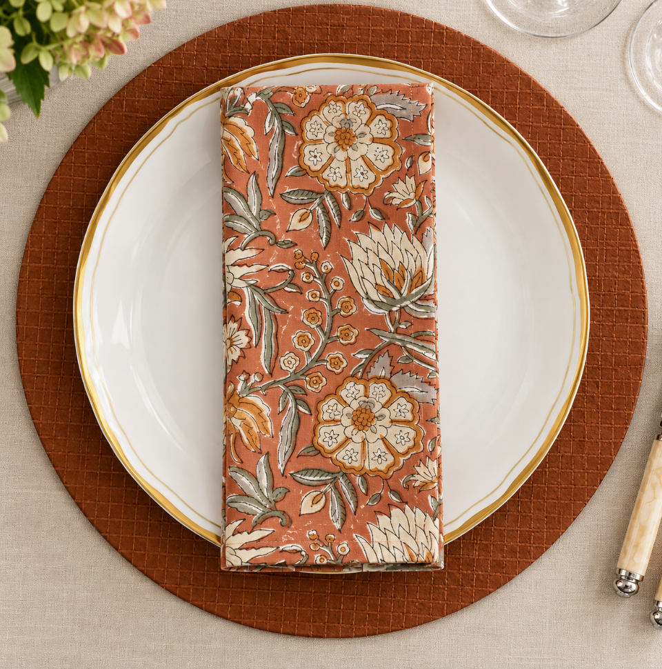 A folded rust-colored Block Print Napkin Set of 4 - Rust with a floral block print pattern resting on a white dinner plate with a gold rim.