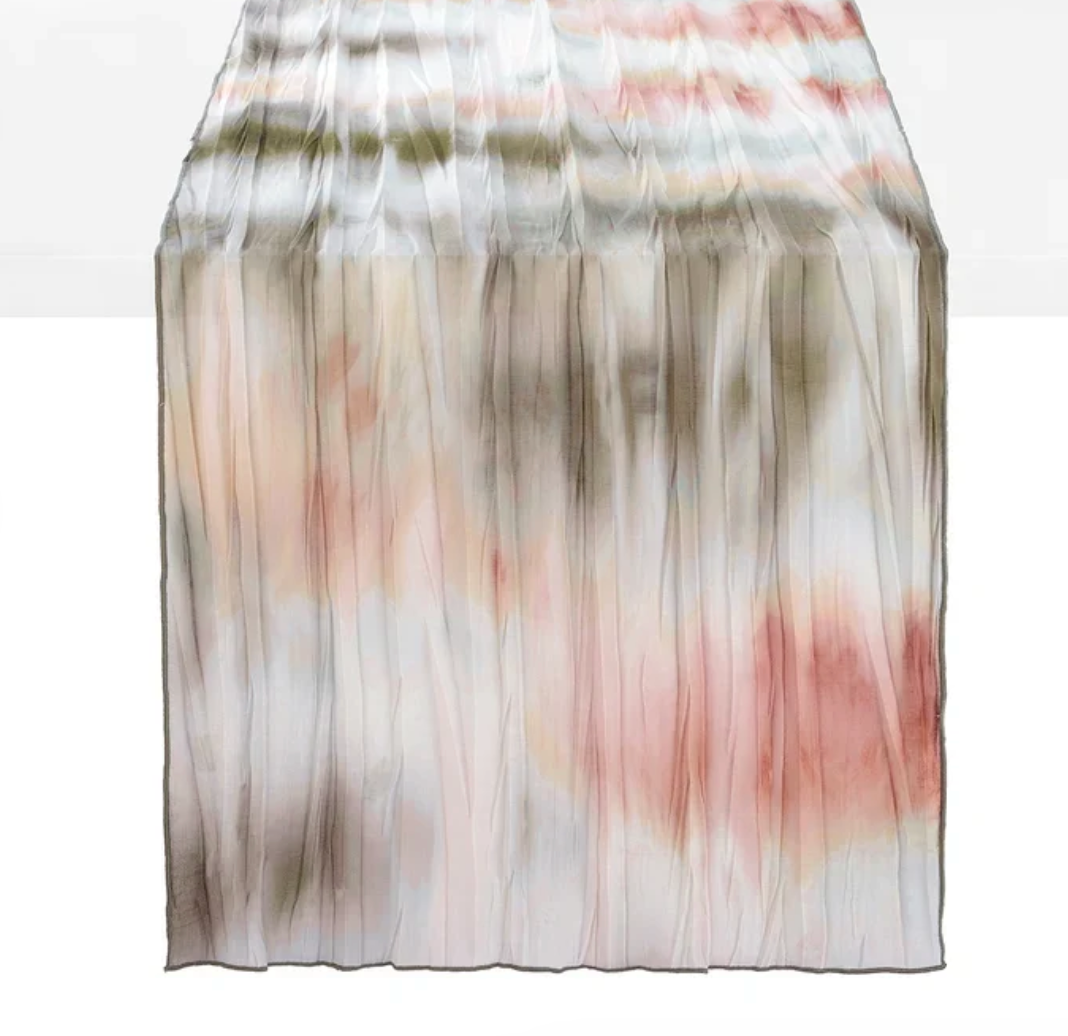 A long, pleated Bodrum Sonata Table Runner 90″ - Woodland featuring a watercolor-style tie-dye pattern in shades of blush, olive green, and gray.