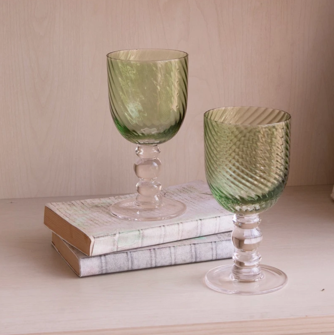 Two green Beatriz Ball GLASS French Swirl Wine glasses with swirling patterns and spherical stems resting on two stacked books.