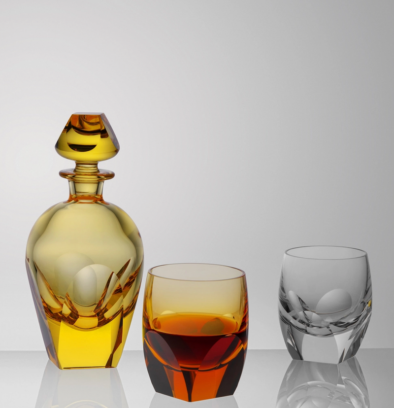 The Moser Bar Decanter 750 ml in yellow with a matching stopper stands beside two short glasses—one filled with amber liquid, the other empty—against a plain light gray background.