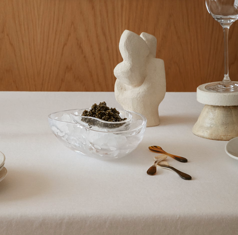 The Georg Jensen Indulgence Caviar Dish rests on a white tablecloth, accompanied by abstract white sculptures, three small spoons, stacked plates, and two empty wine glasses against a wood backdrop.
