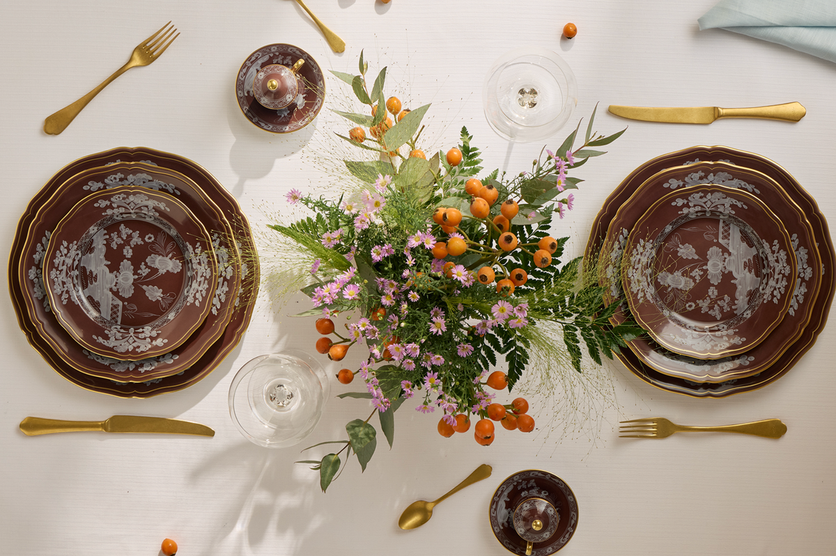 Elegant table set with Ginori 1735's Oriente Italiano Castagna china, brown and white details, gold cutlery, clear glassware, and a centerpiece of orange berries and pink flowers atop a crisp white cloth. Made in Italy.