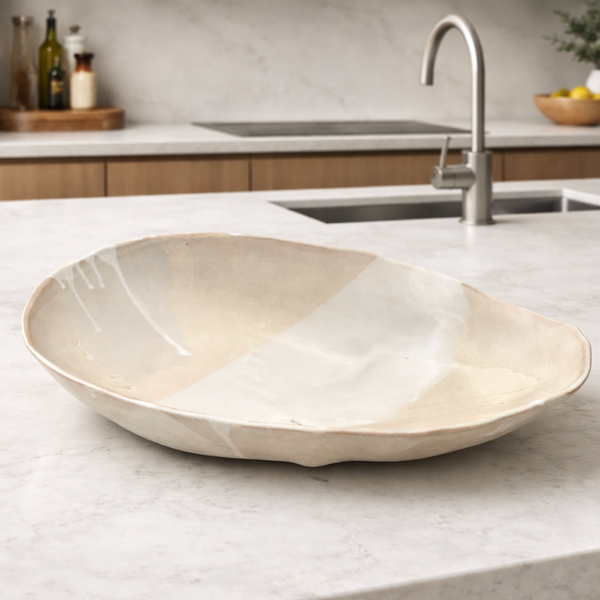 The Ripple Oval Large Bowl by Alex Marshall Studios, a handmade ceramic with a glossy off-white finish, sits on the marble countertop—ideal for serving salad in your modern kitchen with wooden cabinets.
