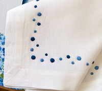 The Bodrum Dots Napkin Set of 4-Blue features white napkins with two lines of blue dots in varying sizes at the corner and a subtle blue floral background, offering an elegant touch to your dining table.