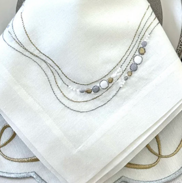 A close-up of Bodrum’s Drift Flow Napkin Gold & Silver Set of 4, crafted from cotton ramie with silver, gold, and grey embroidered curved lines and circular edge patterns.