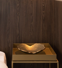 The Mapa Gold & Bronze Trim Bowl by Gardeco sits on a gold side table beside a bed, beautifully contrasted against a dark wood-paneled wall.