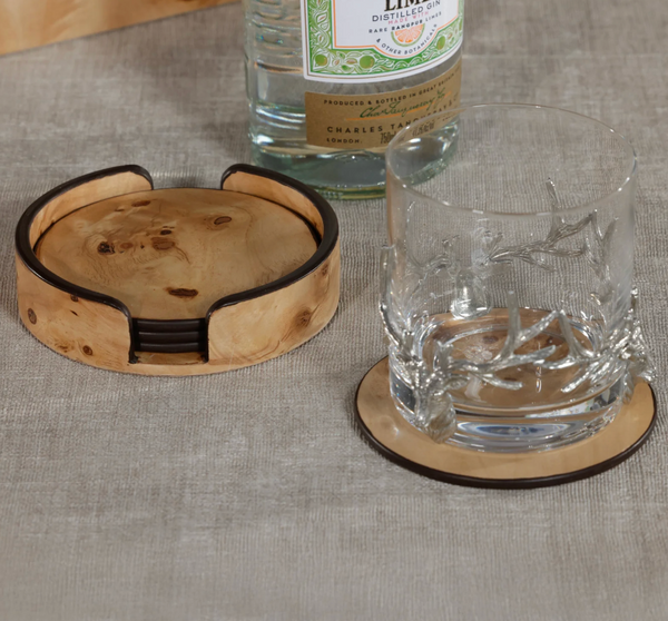A tumbler on a wooden coaster from the Zodax Valari Burl Veneer Coaster Set of 4 sits beside its matching holder and stacked coasters, all arranged on gray fabric with a bottle of distilled gin visible in the background.