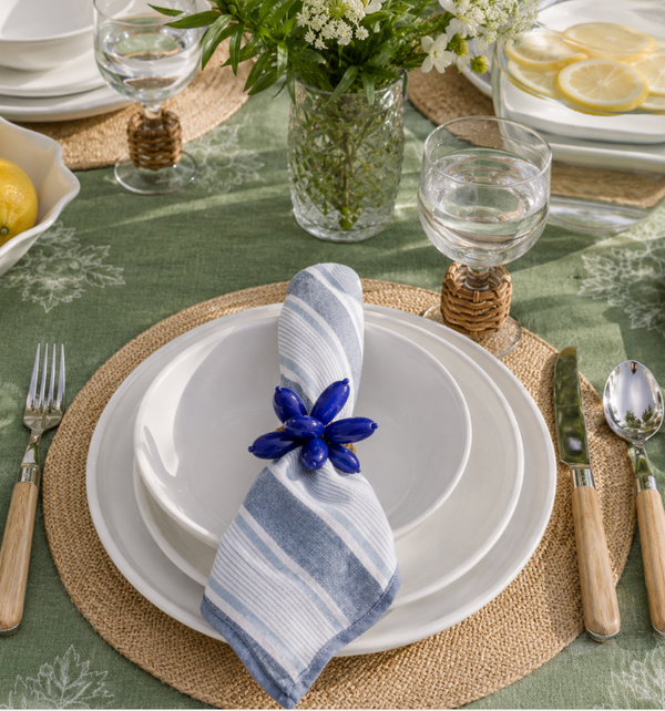 A stylish place setting on a green leaf-patterned tablecloth features Relish Double Line Melamine Dinnerware White (BPA-free), a striped napkin with blue ring, wooden-handled cutlery, water glass, and fresh centerpieces—perfect for outdoor entertaining.