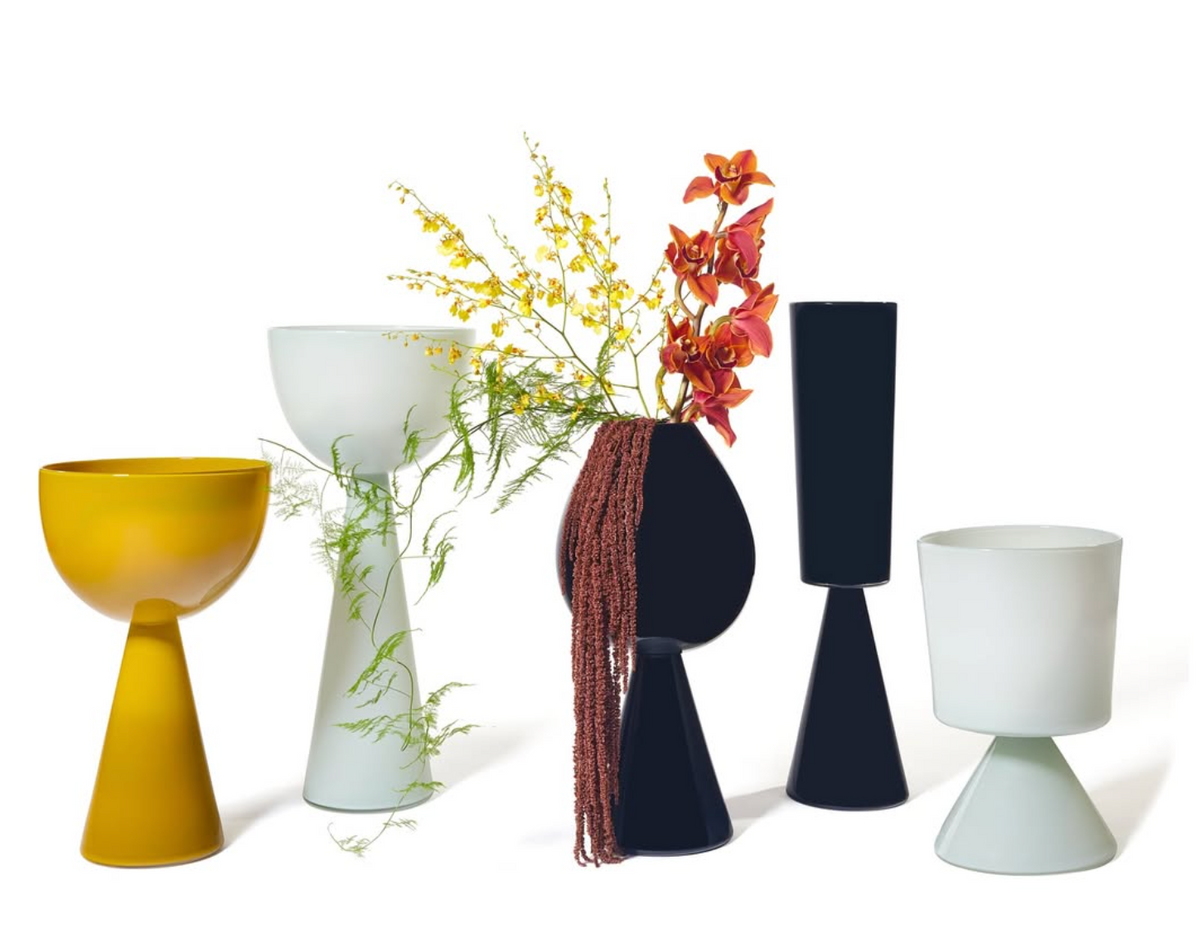 Five modern Fiorelli Vases Ash from Current Home NY—sculptural in yellow, white, and black—showcase yellow and orange flowers with green foliage, creating a striking floral arrangement against a white background.