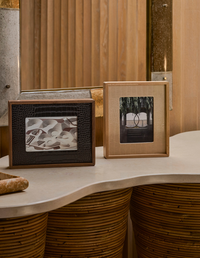 A Terreno Leather & Walnut Frame by Greg Natale and a plain light wood frame sit on a wavy beige countertop with wooden paneling, mirrors, and modern architectural details in the background.