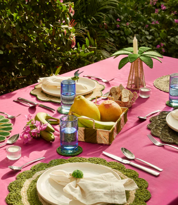 A vibrant outdoor table setting with a pink tablecloth, green placemats, white plates, blue glasses, cutlery, and Coro Cora’s Palm Tree Candle Holder in Navy (Medium), plus fruit basket, bread, and tropical plants in the background.