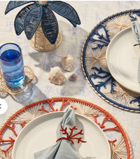 A coastal-themed table setting features two white plates on Iraca palm placemats—one orange, one blue. Each plate has a blue napkin with a coral ring. Nearby are seashells, a palm-shaped decor, and Coro Cora’s navy Palm Tree Candle Holder (Medium).