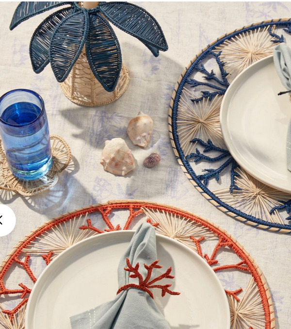A coastal-themed table setting features two white plates on Iraca palm placemats—one orange, one blue. Each plate has a blue napkin with a coral ring. Nearby are seashells, a palm-shaped decor, and Coro Cora’s navy Palm Tree Candle Holder (Medium).