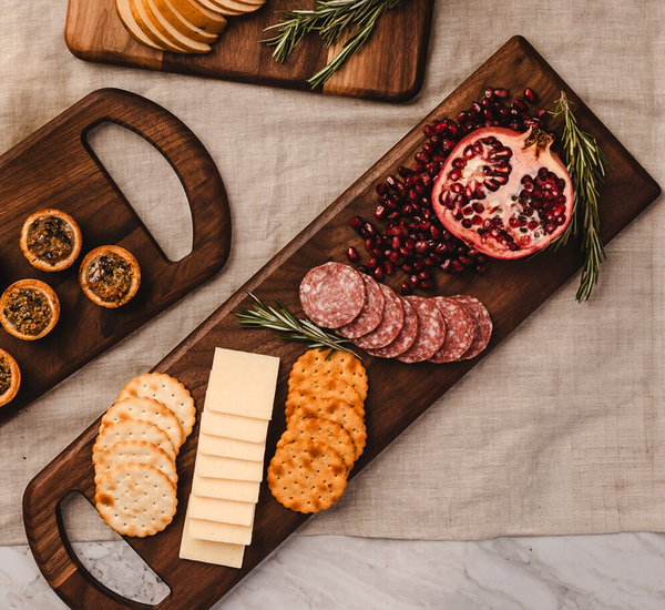 A Bristal Walnut Serving Board by JK Adams holds crackers, cheese slices, salami, halved pomegranate, and rosemary sprigs. Another tray displays crackers and small tarts—all elegantly set out on a neutral tablecloth.