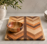 A small wooden bowl sits on the Multiwood Herringbone Serving Board by Dickinson Woodworking, with a plant in a ceramic vase and stacked white plates in the background on a marble countertop.