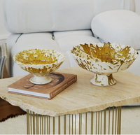 Two Jagged Edge Pedestal Fruit Bowls in Ochre Splatter from Moroccan White rest on a light stone coffee table—one bowl is artfully placed atop a closed book, while the other sits directly on the table surface. A white sofa provides a soft backdrop.