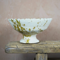 The Jagged Edge Pedestal Fruit Bowl in Ochre Splatter by Moroccan White—a handmade, white ceramic bowl featuring a scalloped edge and distinctive ochre splatter design—sits atop a rustic wooden surface against a pinkish textured wall, evoking the artistry of traditional Moroccan craftsmanship.