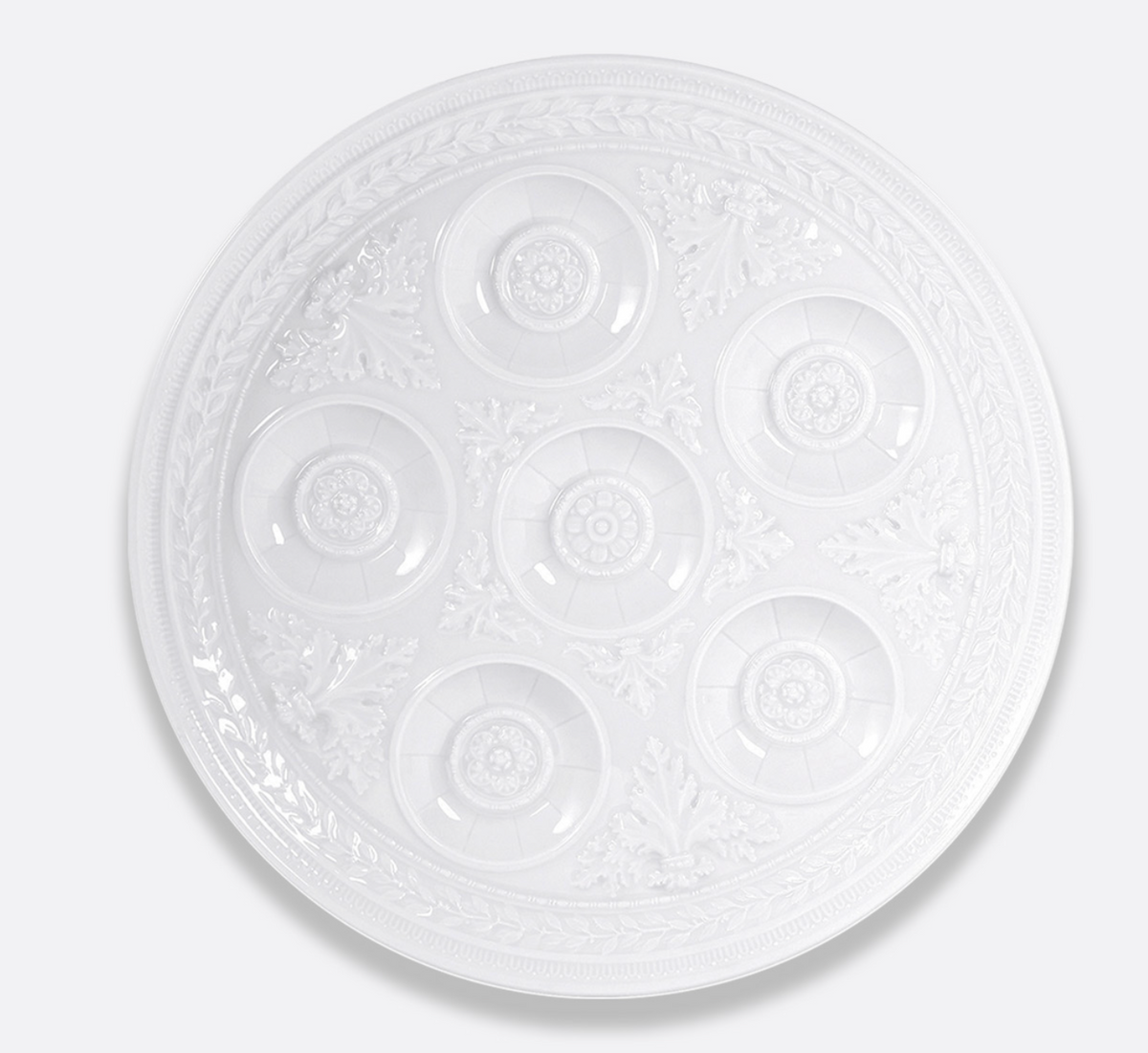 Introducing the Bernardaud Louvre Dinnerware Seder plate—a white, round piece featuring seven decorative, indented sections arranged in a circle and adorned with ornate floral patterns around the edges, inspired by the elegance of French architecture.