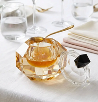 A faceted Crystal Honey Jar w/ Spoon in Amber / Black by BT- Shalom, filled with golden honey, sits with its lid off on a white tablecloth beside clear glassware and folded napkins—perfect for adorning your Rosh Hashanah Yom Tov table. The elegant jar is accompanied by a matching gold spoon.