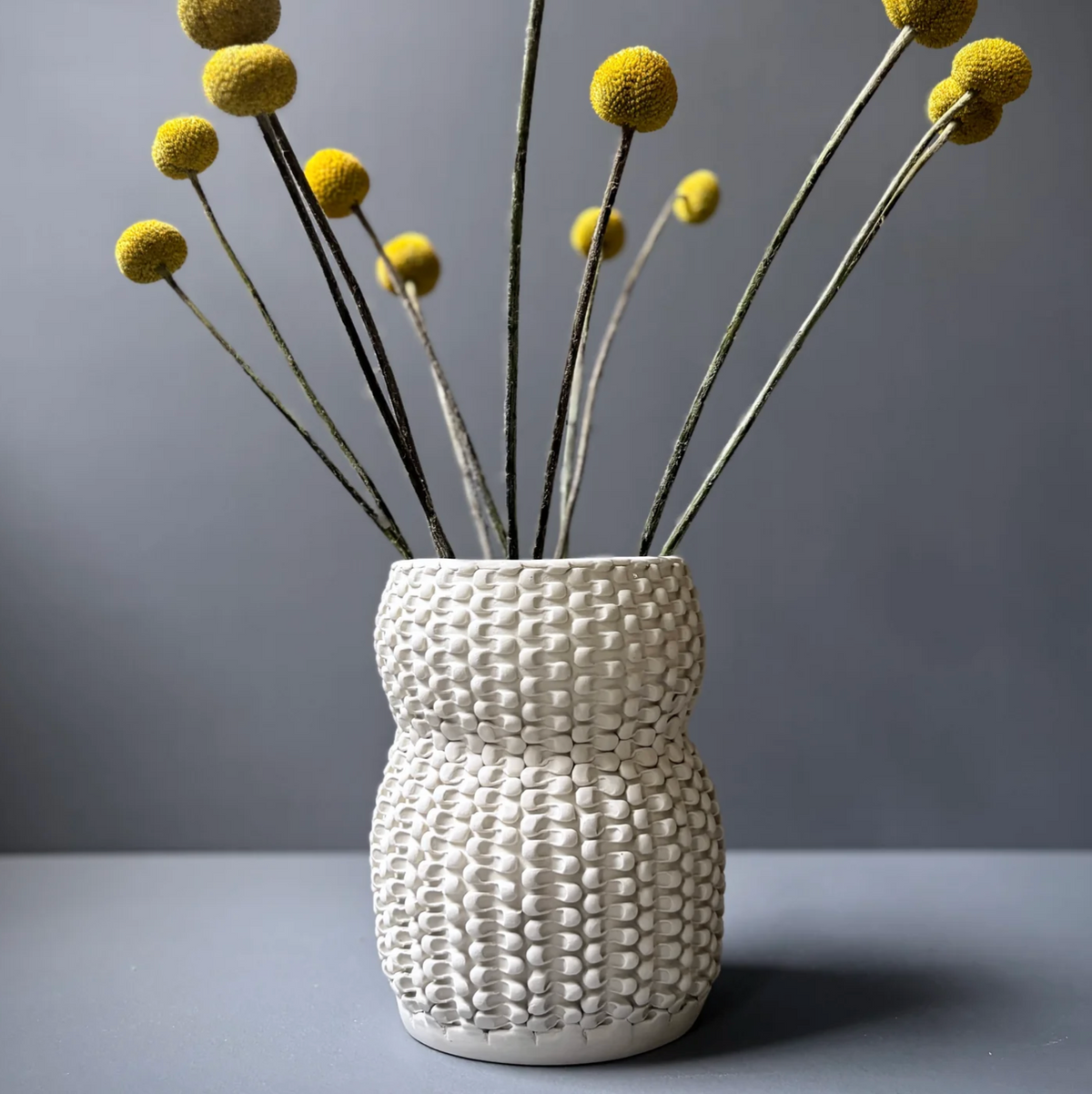 A cream-colored Chain Curve Vase from CYM Warkov Ceramics features a textured, woven pattern and holds long stems with round yellow billy ball flowers, creating a striking decorative accent against a plain gray background.