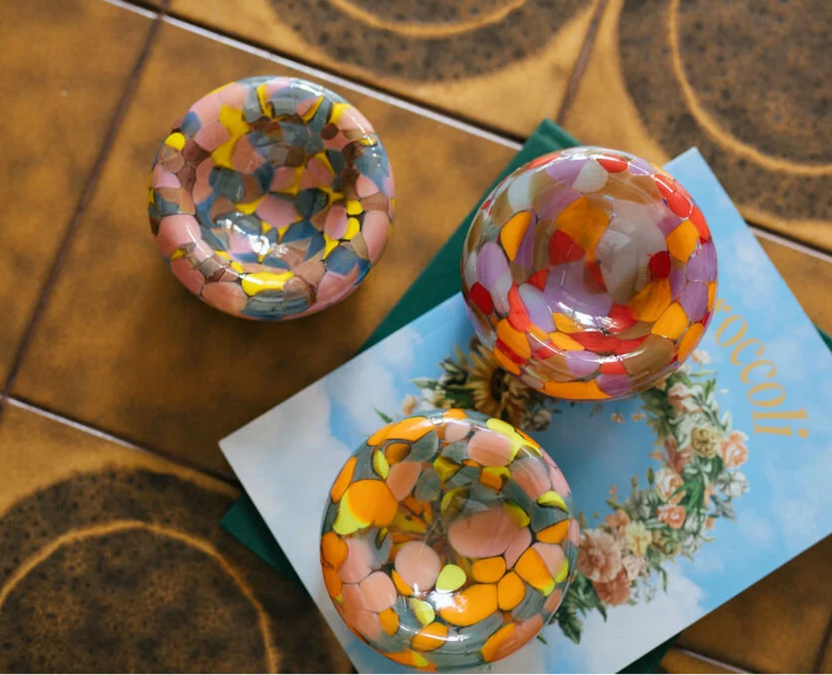 Three colorful, round Maria Ida Designs Jelly Baby Bubble Bowls with mosaic patterns sit on a patterned brown surface next to a book with a floral wreath on the cover.