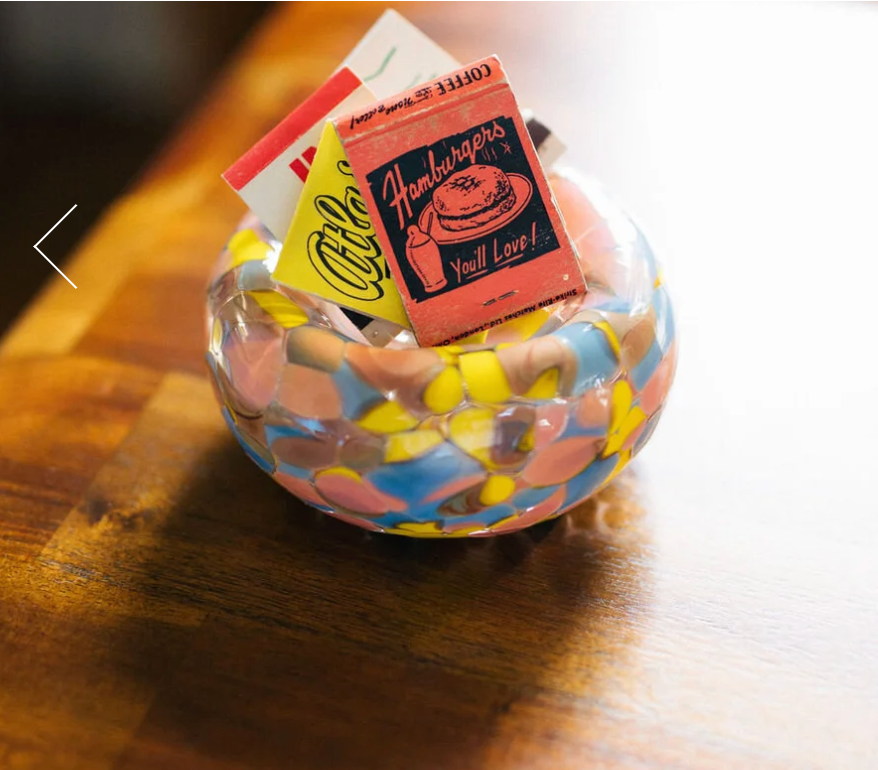 A colorful Jelly Baby Bubble Bowl by Maria Ida Designs holds vintage matchbooks—including ones advertising hamburgers and a café—on a wooden surface. This striking piece of art glass brings vibrant character to the scene.
