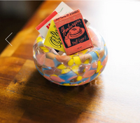 A colorful Jelly Baby Bubble Bowl by Maria Ida Designs holds vintage matchbooks—including ones advertising hamburgers and a café—on a wooden surface. This striking piece of art glass brings vibrant character to the scene.