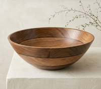 The Volare Large Serving Bowl by Be Home, crafted from round acacia wood, rests on a light-colored surface with a minimal background and a sprig of greenery in the corner.