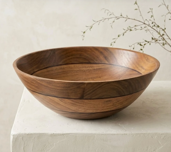 The Volare Large Serving Bowl by Be Home, crafted from round acacia wood, rests on a light-colored surface with a minimal background and a sprig of greenery in the corner.