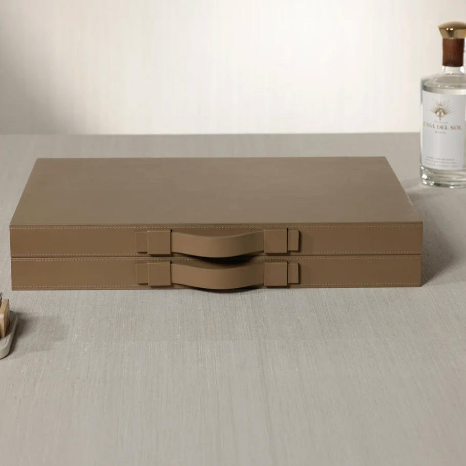 Two stacked taupe Gene Leather Backgammon cases by Zodax, each featuring a sleek handle, rest atop a light gray tabletop—a subtle nod to the refined elegance of this handcrafted set. In the background, a bottle of clear liquid and a small dish enhance the sophisticated ambiance, creating an atmosphere perfect for enjoying a luxury game board experience.