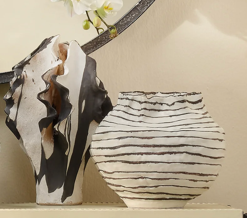 Two decorative ceramic vases sit side by side. One is the Jamie Young Cocoon Vase Wide, featuring an abstract, wavy black-and-white design with sculpted curves, while the other is a round vase with irregular horizontal black stripes on white. Part of a white orchid appears above.