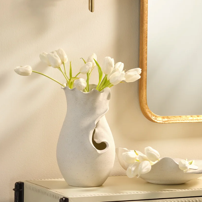 White tulips in the Jamie Young Fabio Vase sit on a cream table next to a matching bowl with tulip petals, beside a gold-framed mirror on a light wall.