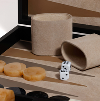 A close-up of a Zodax Gene Leather Backgammon - Midnight Black set with tan and black pieces, two white dice showing five and six, and two beige dice cups, one upright and one on its side.