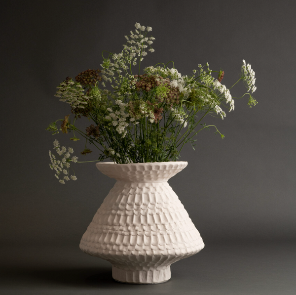 The Teramo Vase by FlowDecor Inc. is a white ceramic piece with a scalloped texture, wide base, and narrow opening, shown filled with white wildflowers and green stems against a dark gray background.