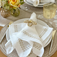 A white linen napkin from the Pearls Napkin Set of 4 - Beige, decorated with a beige beaded border and held by a jeweled bee napkin ring on a white plate.