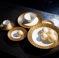 Two L’Objet Aegean Gold - Tea Cup and Saucer (Set of 2) porcelain tea cups with 24k gold handles and three white plates with textured gold rims, arranged on a dark surface with two pieces of fruit.