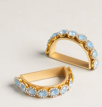 A set of four Amaryllis Golden Bar Napkin Ring elegant, gold-toned semi-circular napkin rings decorated with rows of light blue stones and clear crystals.