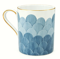 Introducing the Marie Daâge Cercle D’Ecailles Gold Filet mug by Marie Daâge—a white ceramic piece adorned with a gold-rimmed handle and top edge, featuring an overlapping blue scalloped pattern reminiscent of fish scales or gentle waves. Inspired by classic French tableware, this mug captures the timeless elegance of Limoges porcelain.