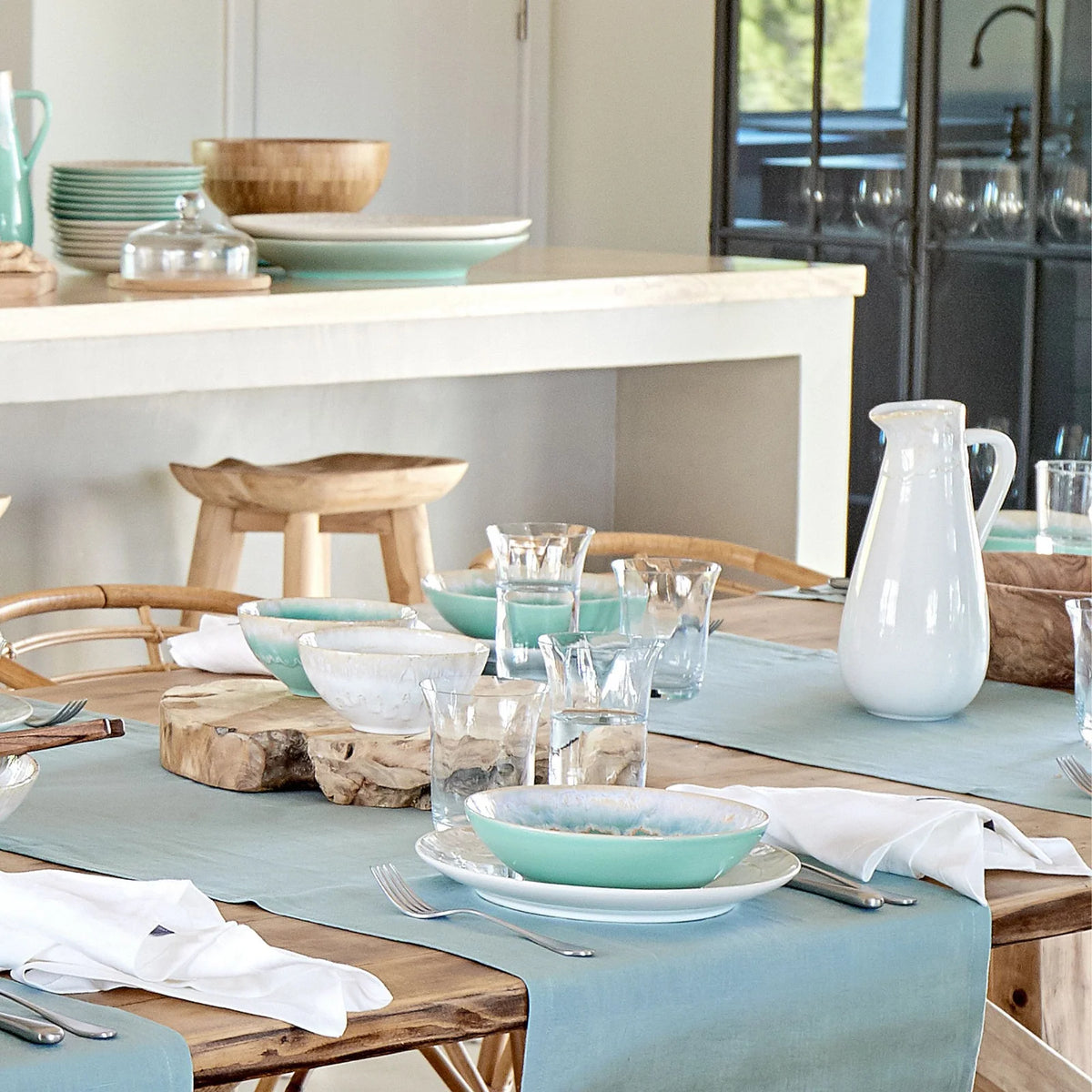 A wooden dining table set with Taormina Aqua Dinnerware, including aqua-rimmed bowls, white plates, glass tumblers, white napkins, and a white pitcher arranged on a light blue table runner.