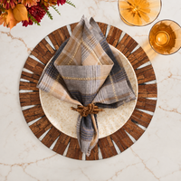 A plate rests on the Halo Placemat in Brown and White by Kim Seybert, adorned with a plaid napkin tied in a decorative knot and flanked by two amber glasses, all arranged on a marble table with flowers in the top corner.