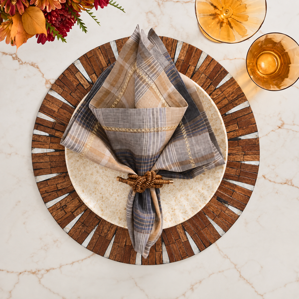 A plate rests on the Halo Placemat in Brown and White by Kim Seybert, adorned with a plaid napkin tied in a decorative knot and flanked by two amber glasses, all arranged on a marble table with flowers in the top corner.