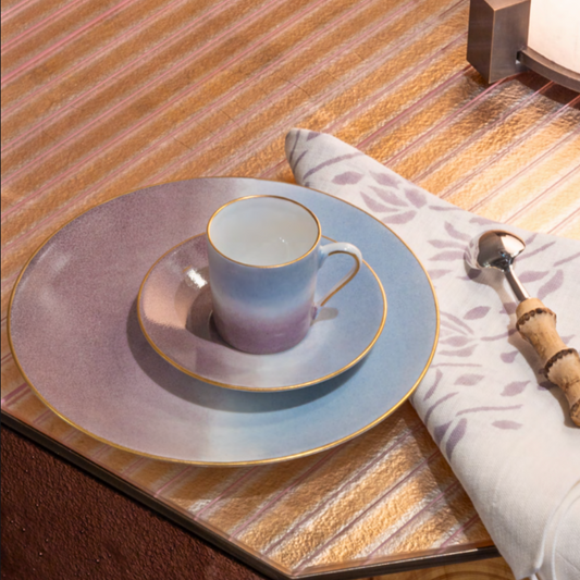 A Marie Daâge Tie Dye Platinum cup and saucer, both hand-painted, rest on a matching dinner plate with platinum rims by Marie Daâge, beside a floral napkin and spoon atop a striped tabletop.