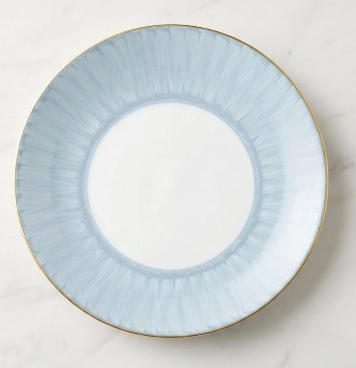 The Marie Daâge Ruban 1 Dinnerware by Marie Daâge is a round Limoges porcelain plate with a wide pale blue rim featuring subtle radial brushstrokes and a thin gold edge, set on light marble. The center remains plain white.