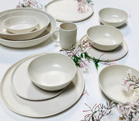 A set of Urban Dinnerware Matte White plates, bowls, and a mug from Alex Marshall Studios are arranged on a white surface, accented by delicate pink and white flower sprigs scattered around them.
