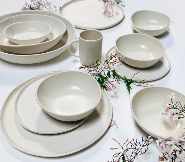 A set of Urban Dinnerware Matte White plates, bowls, and a mug from Alex Marshall Studios are arranged on a white surface, accented by delicate pink and white flower sprigs scattered around them.